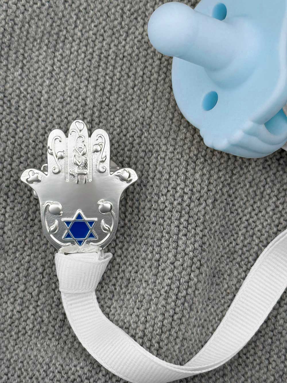 Silver Hamsa charm with Star of David on a white ribbon, next to a blue pacifier on a textured gray background.
